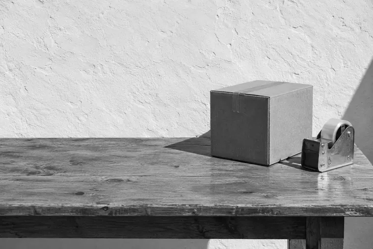 Un carton d’expédition scotché et un dévidoir de ruban adhésif posés sur une table nue, en plein soleil.