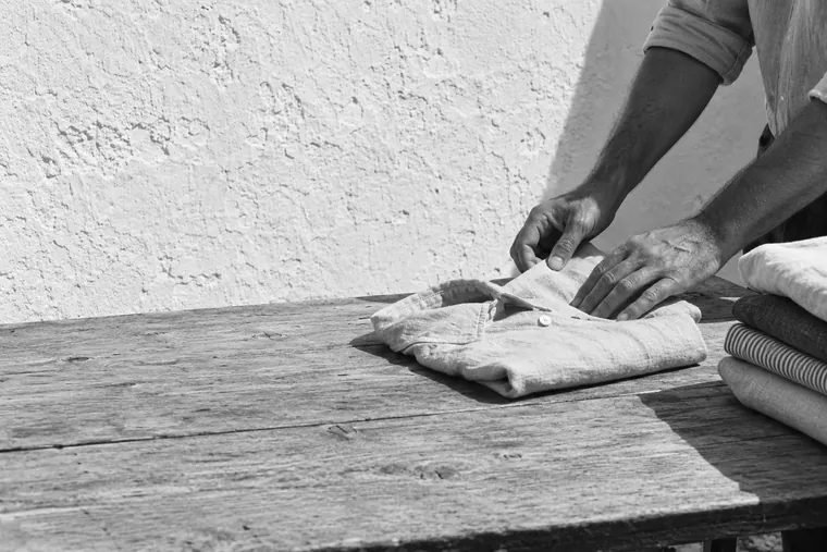 Deux mains plient une chemise de lin sur une table de bois nue, une pile de vêtements pressés au bord du cadre, mur chaulé en plein soleil.