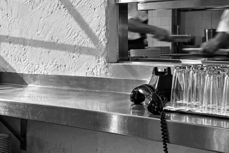 Le passe d’un restaurant en plein service : des verres alignés au premier plan, un combiné téléphonique décroché posé sur l’inox.